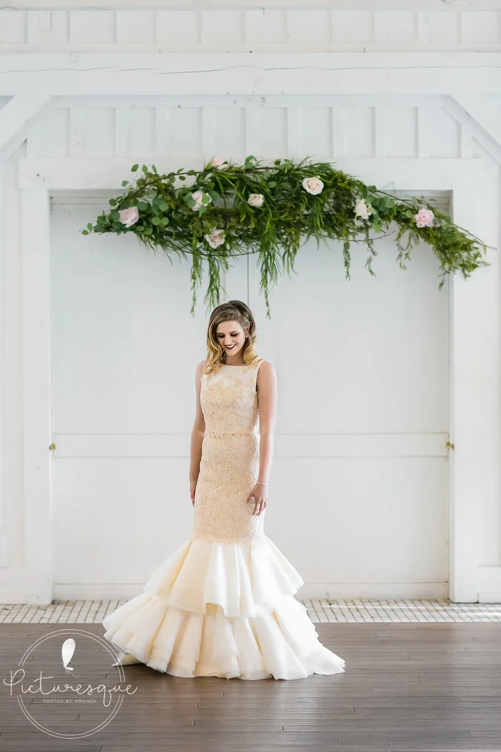 A bride in a gold lace fit-and-flare gown with a tiered organza skirt beneath a floral arch